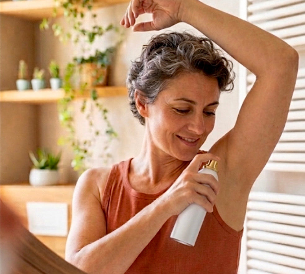 Lächelnde Frau im rostbraunen Top benutzt ein Deodorant-Spray in einem hellen Badezimmer vor einem Spiegel.